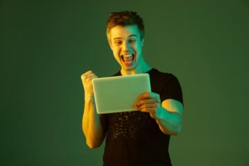 holding tablet celebrating win bet game caucasian man s portrait green studio background neon light beautiful male model concept human emotions facial expression sales ad